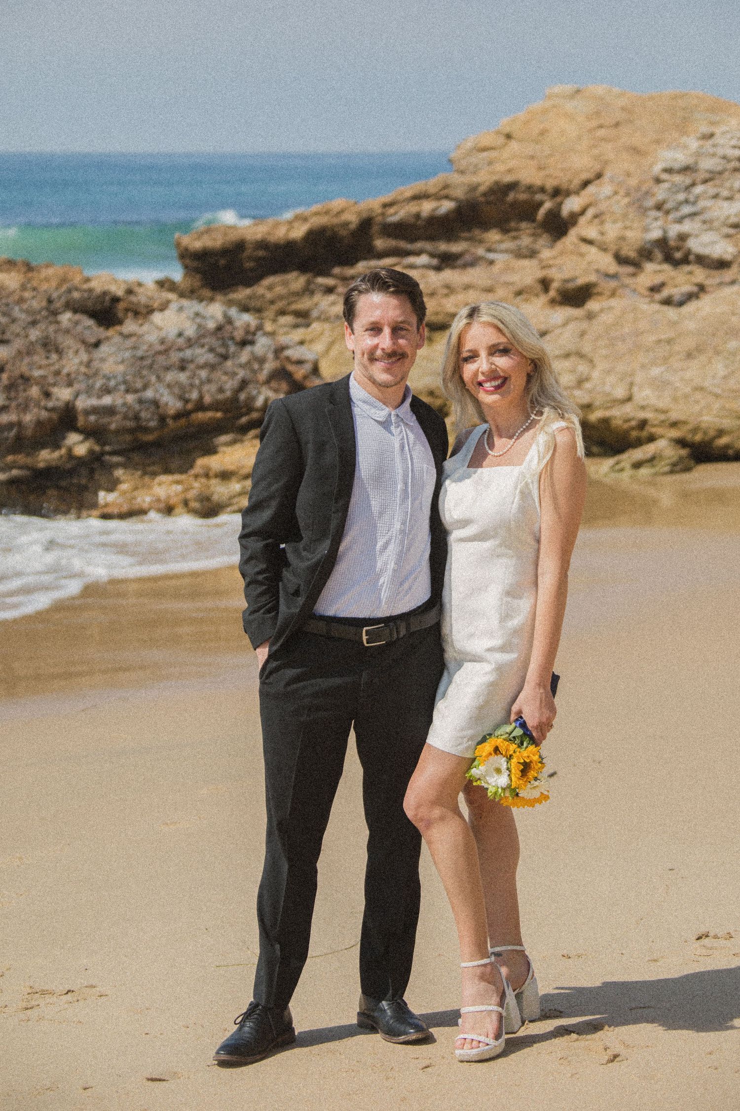 A couple poses on a scenic beach with rocky cliffs in the background during their wedding celebration.