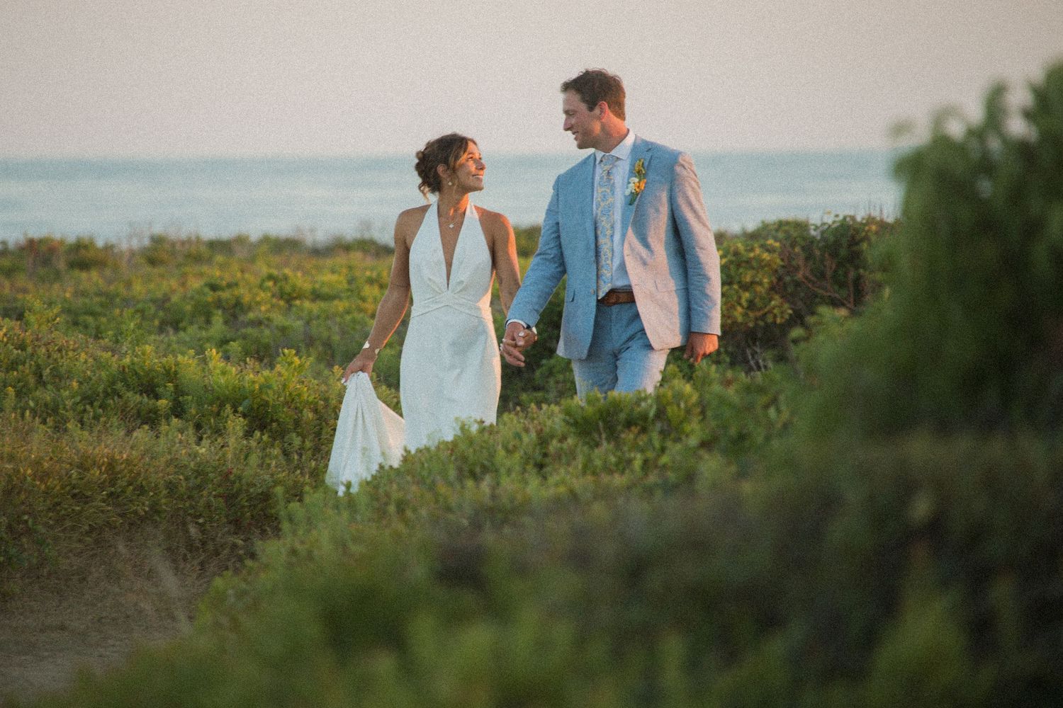 A couple walks through coastal vegetation with ocean views during golden hour.