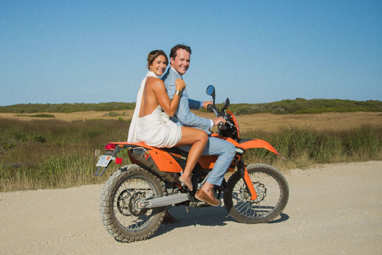 Wedding couple riding together on orange dirt bike along coastal path.