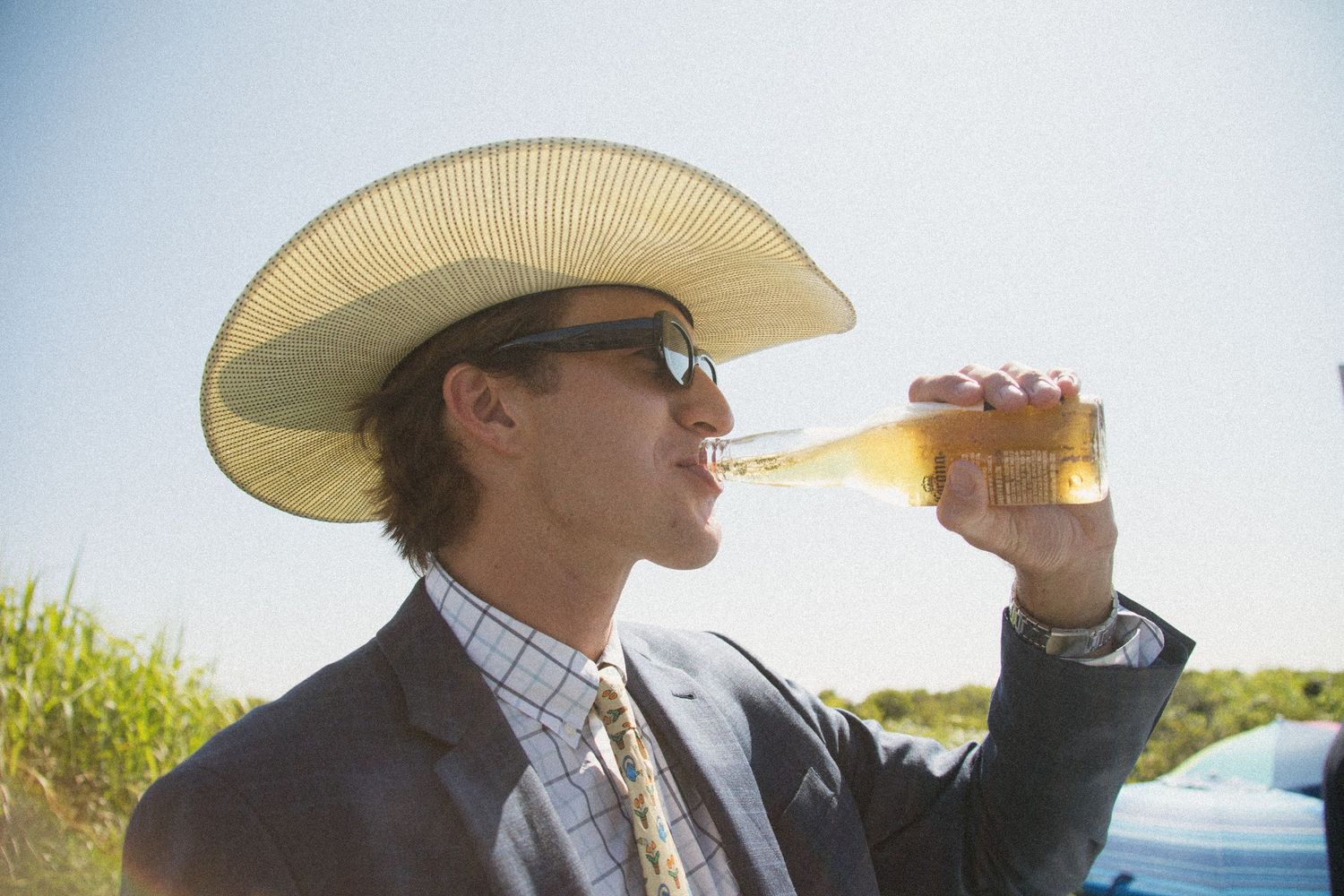 Figure in sunhat and suit drinking from bottle against sunny outdoor backdrop.