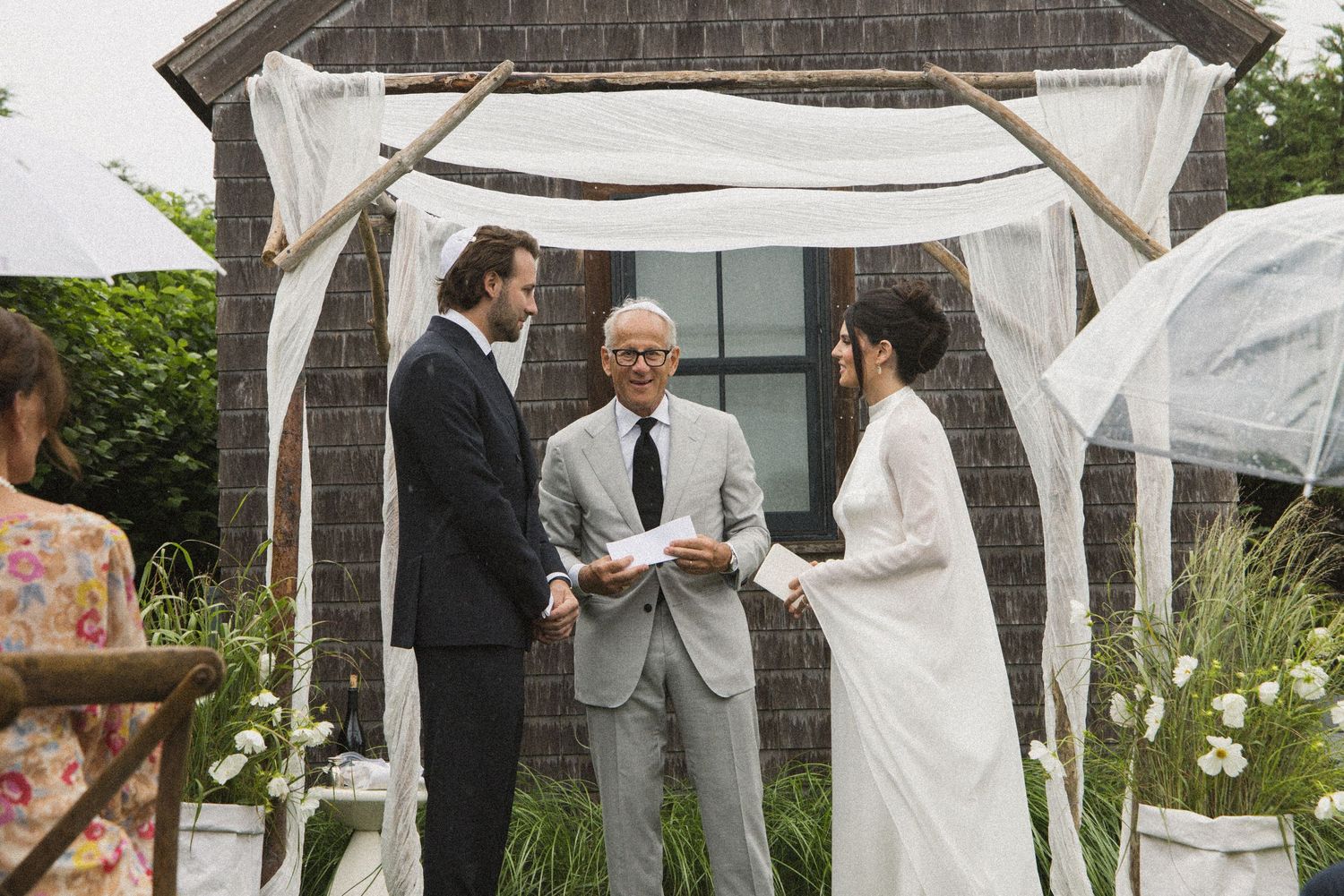 Outdoor wedding ceremony with couple exchanging vows under a rustic arch draped in white fabric, officiated by an elderly man.