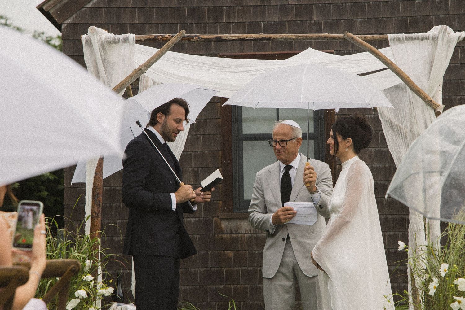 Outdoor wedding ceremony with bride and groom under a rustic wooden chuppah, officiant reading vows, guests holding white umbrellas.