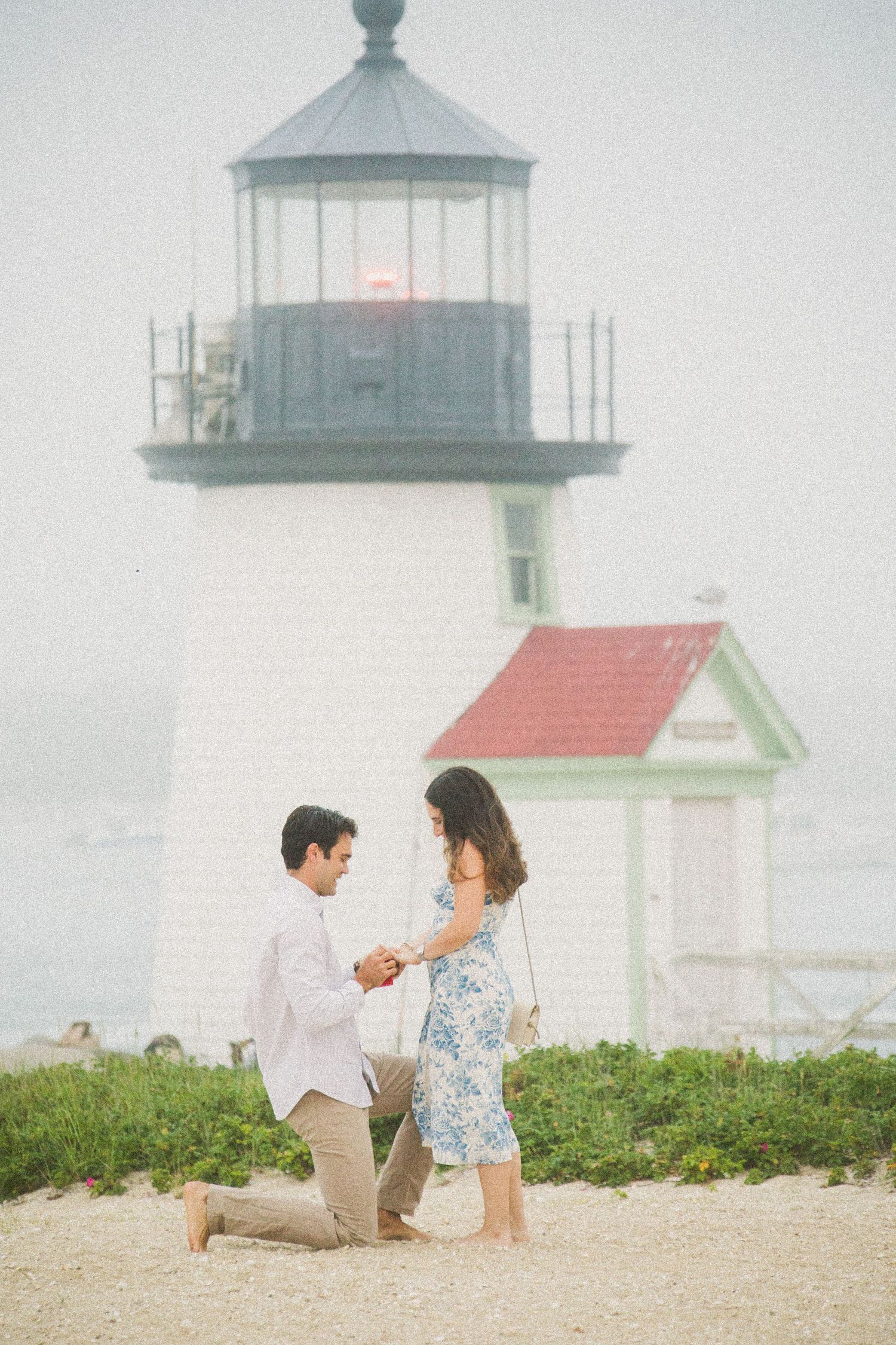 Man proposes to woman on sandy beach with historic lighthouse and red-roofed keeper's cottage in misty background.