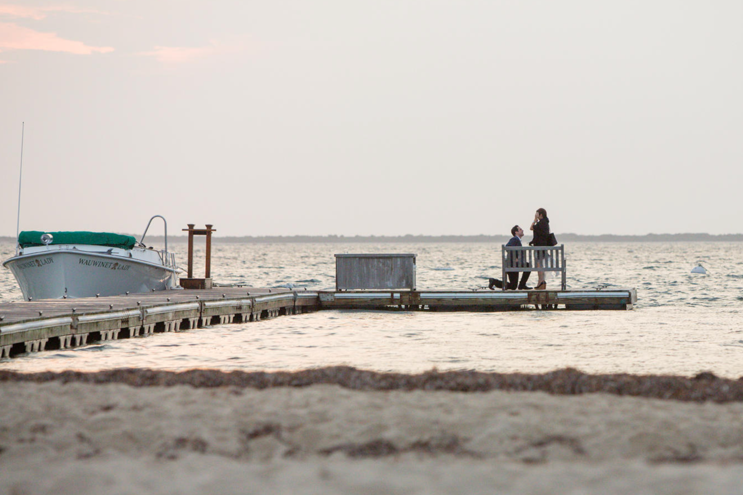 Two people sit on a dock at sunset beside a moored boat on a calm sandy beach shoreline.
