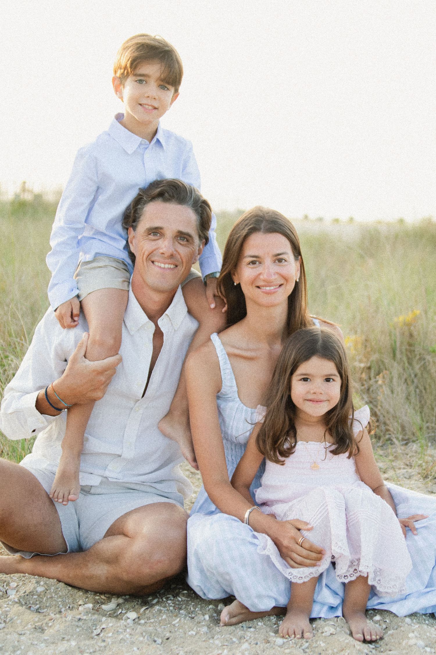 Family dressed in light colors sitting together on a sandy beach at sunset.