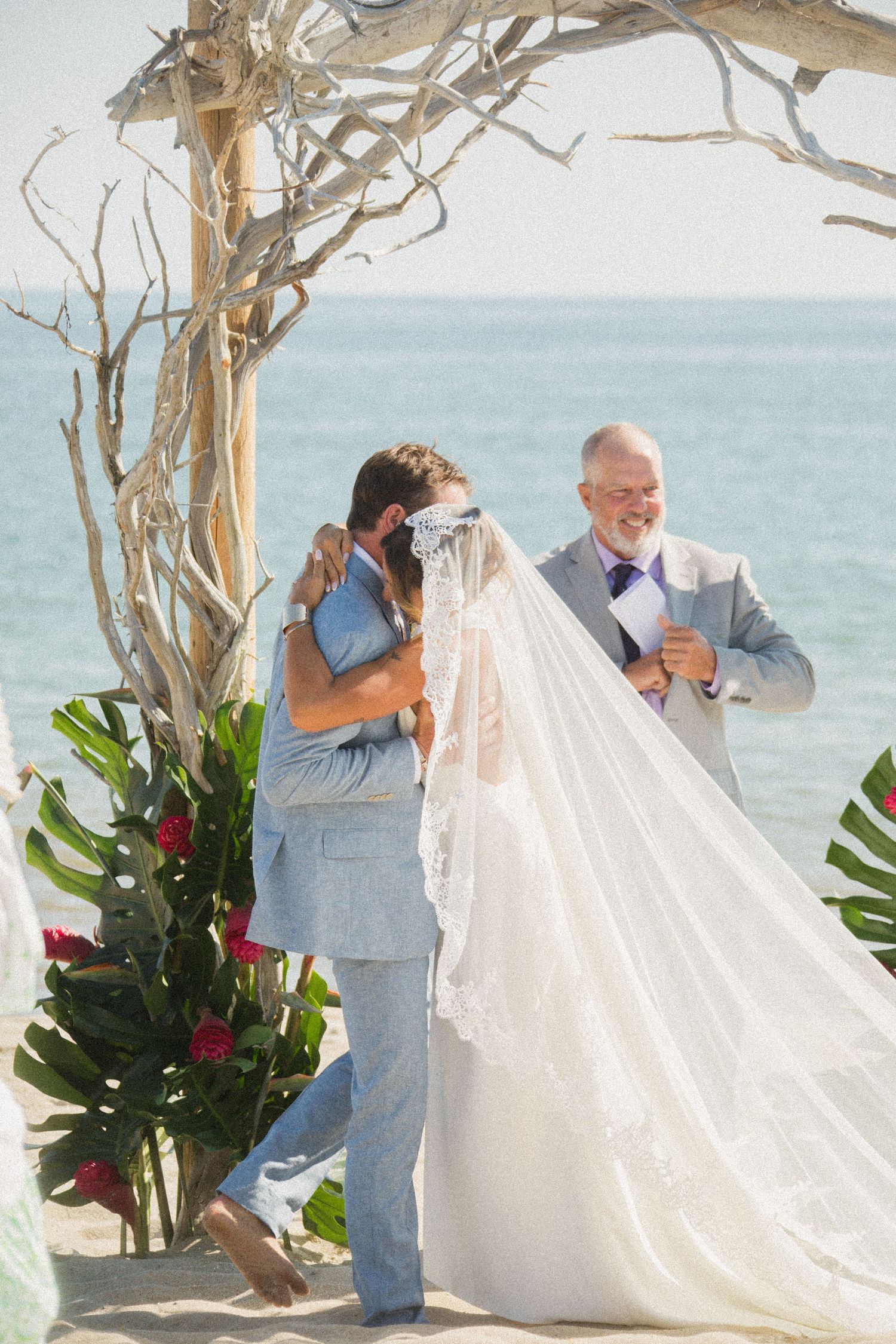 Couple sharing romantic moment under driftwood arch during beach ceremony.