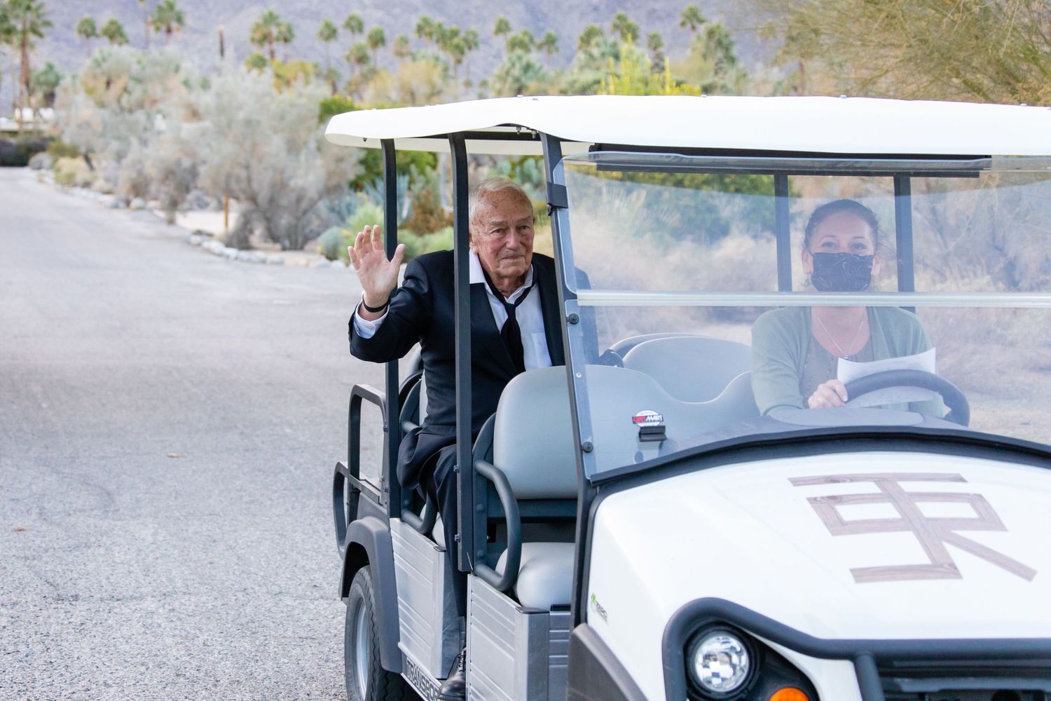 A white golf cart drives along a desert path with mountains visible in the distance.