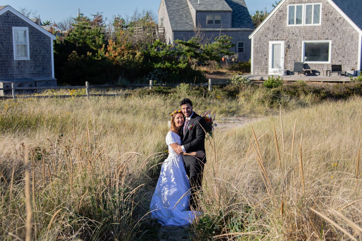Coastal wedding portraits with beach grass and Cape Cod style houses in the background.