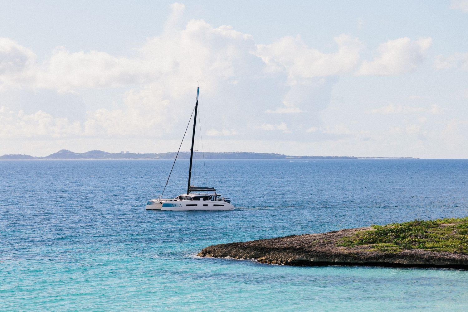 Catamaran sailboat anchored in turquoise waters near rocky coastline.