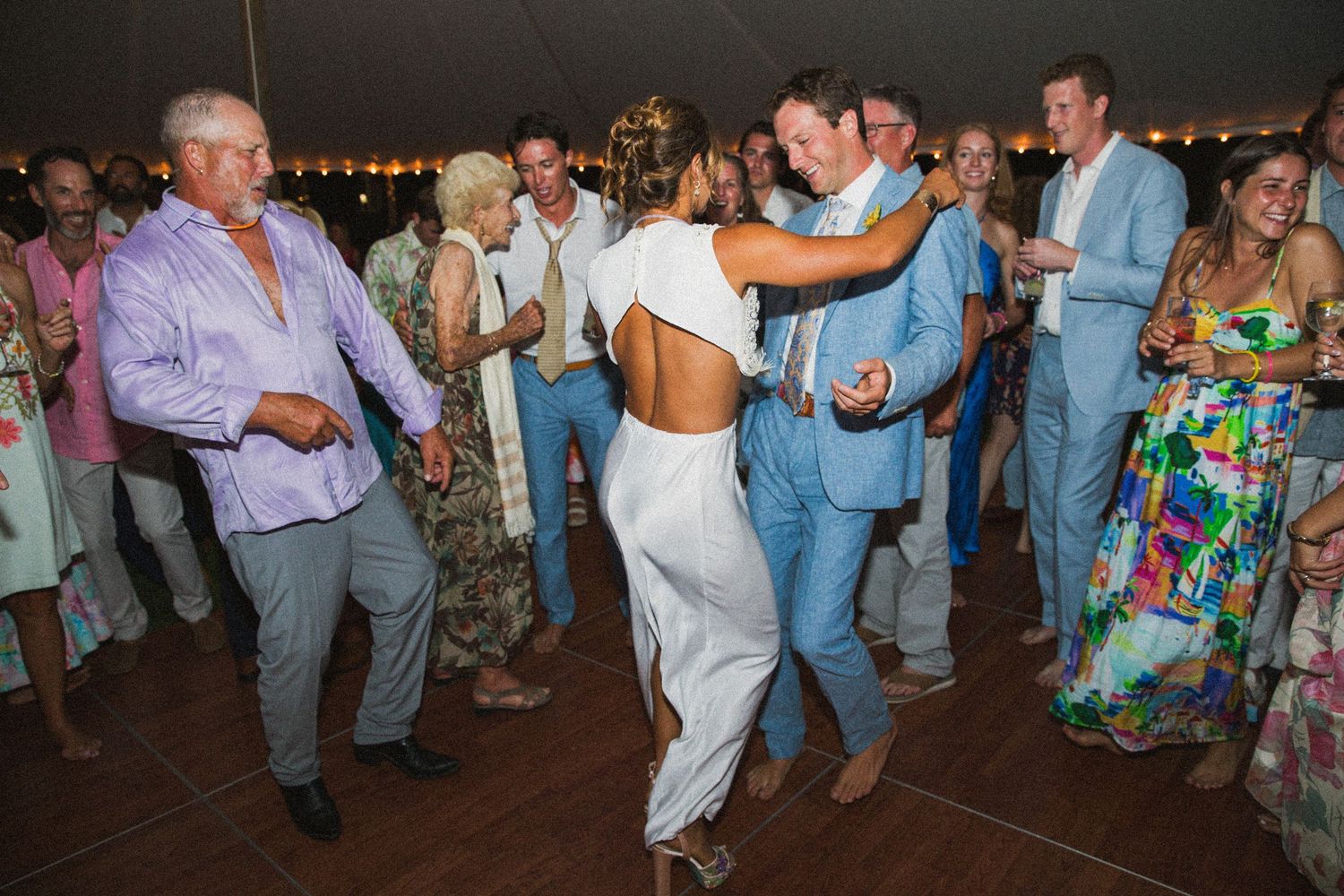 Wedding guests dancing in formal attire under a large white tent during evening reception.