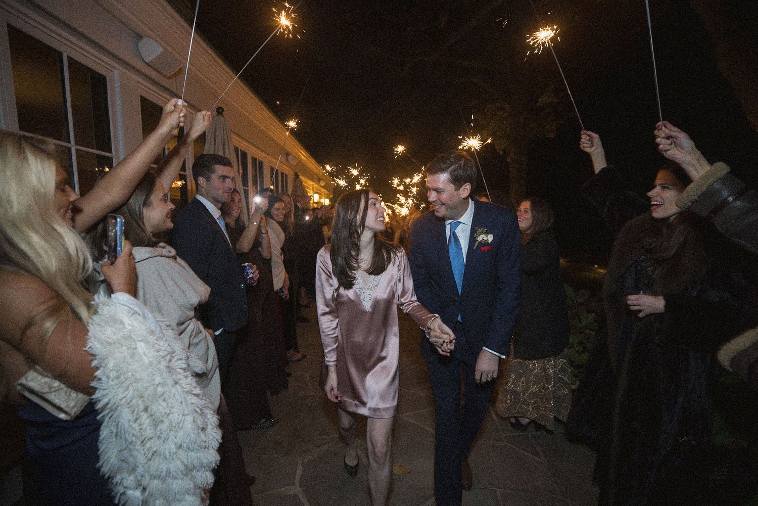 A couple holds hands and walks through a sparkler send-off as guests line the path at a night wedding.
