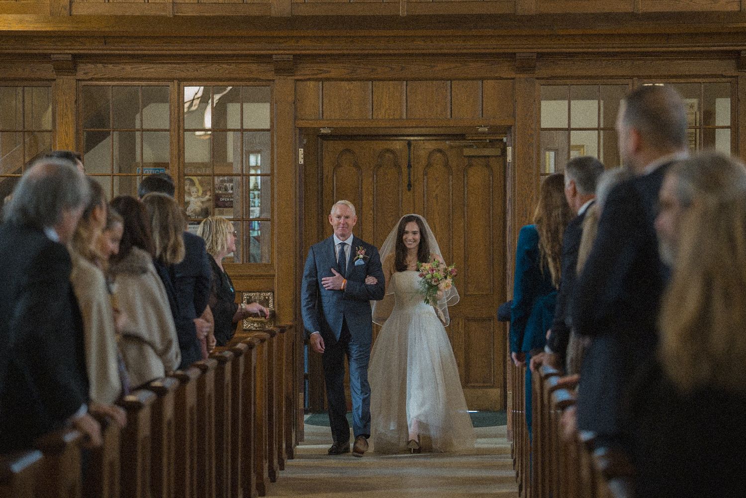 Bride in a white gown and veil walks down the church aisle on her father's arm as seated wedding guests look on.