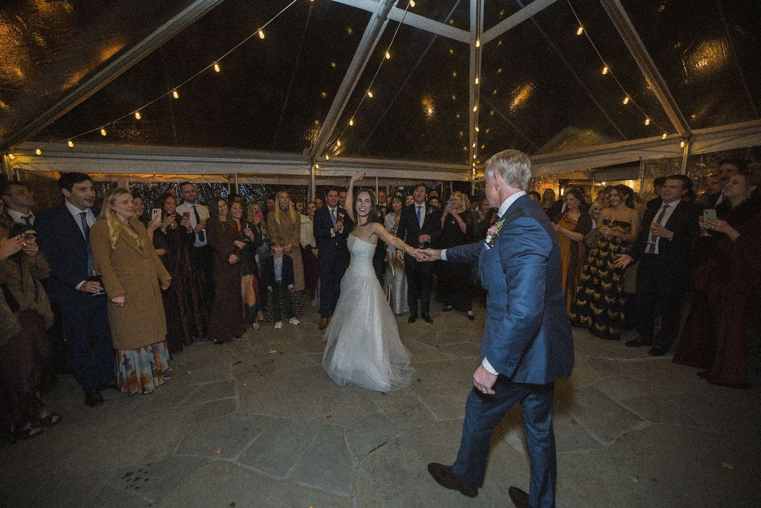 Bride in a white gown and groom in a navy suit share a first dance as wedding guests watch under a tented reception with string lights.