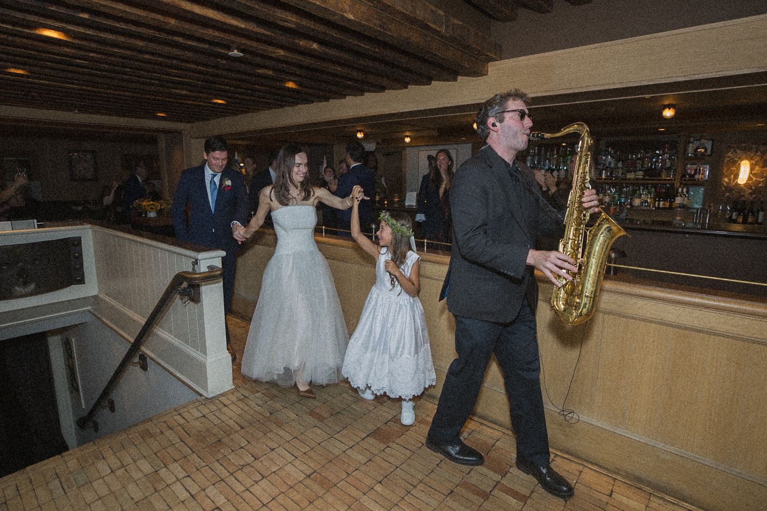 Saxophonist leads a bride in a white gown and a flower girl across a rustic wedding reception venue with wood beams and a bar.