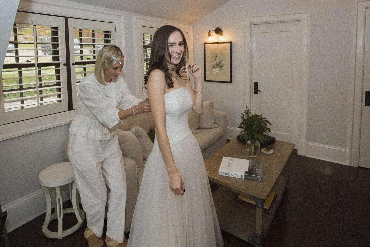 A bride in a strapless white wedding gown smiles while a woman in white helps fasten the back in a bright, cozy room.