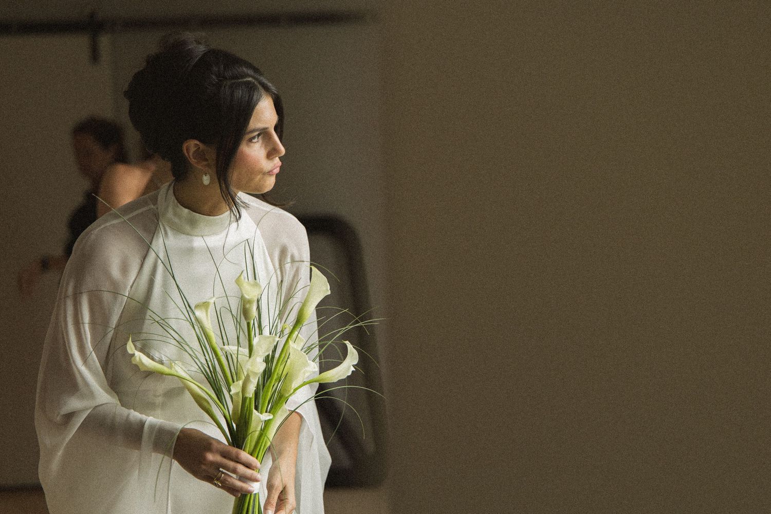 Woman in white dress holding calla lilies bouquet, standing elegantly in soft natural light.