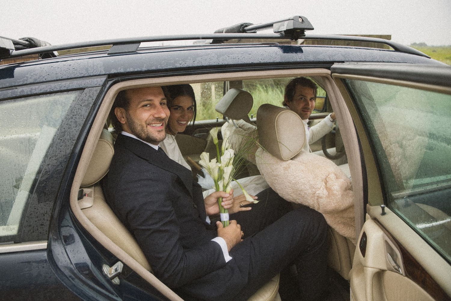 Smiling groom in suit sits in luxury SUV holding white flowers, with wedding party visible in background.