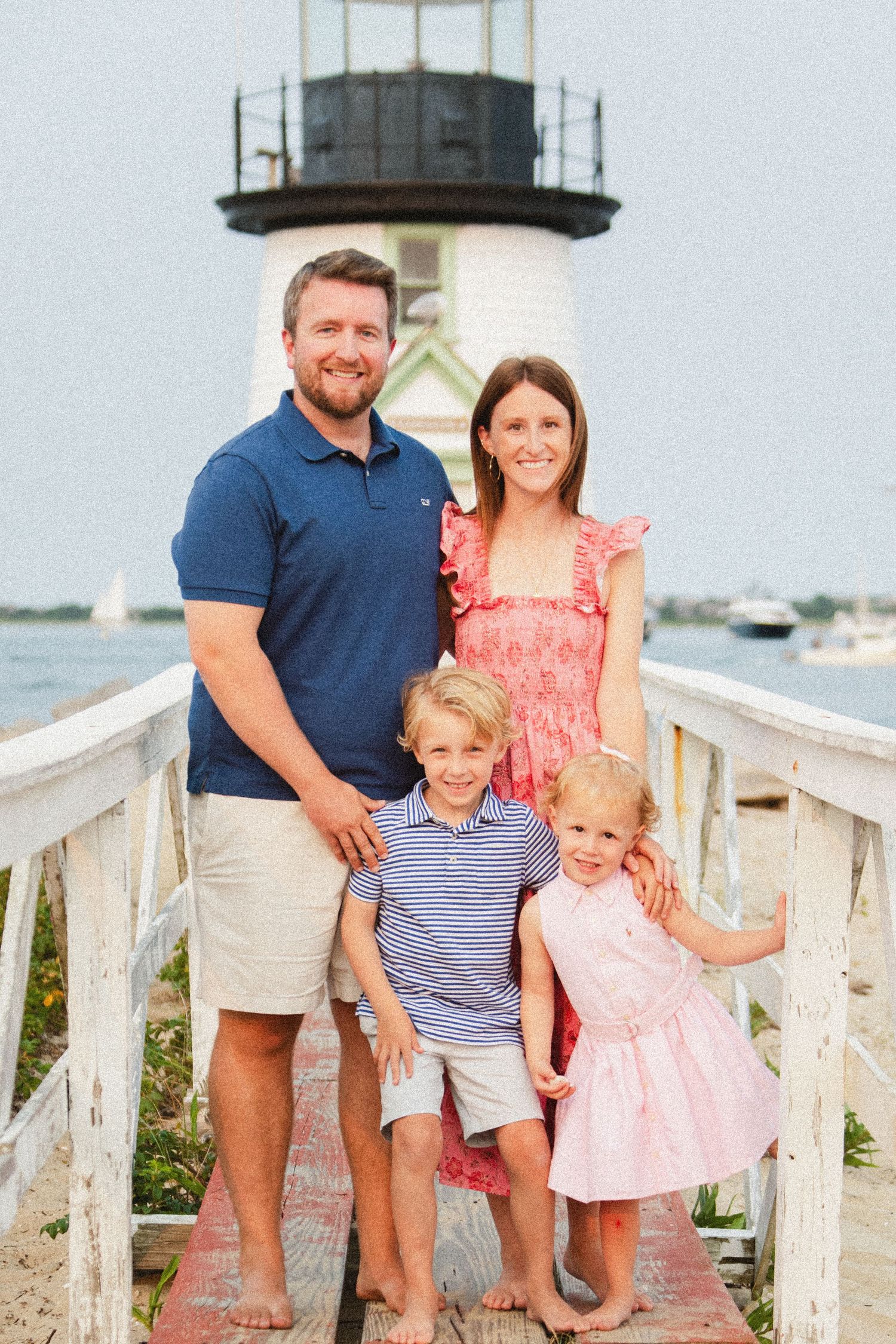 A family poses together on a lighthouse pier with boats in the harbor behind them.