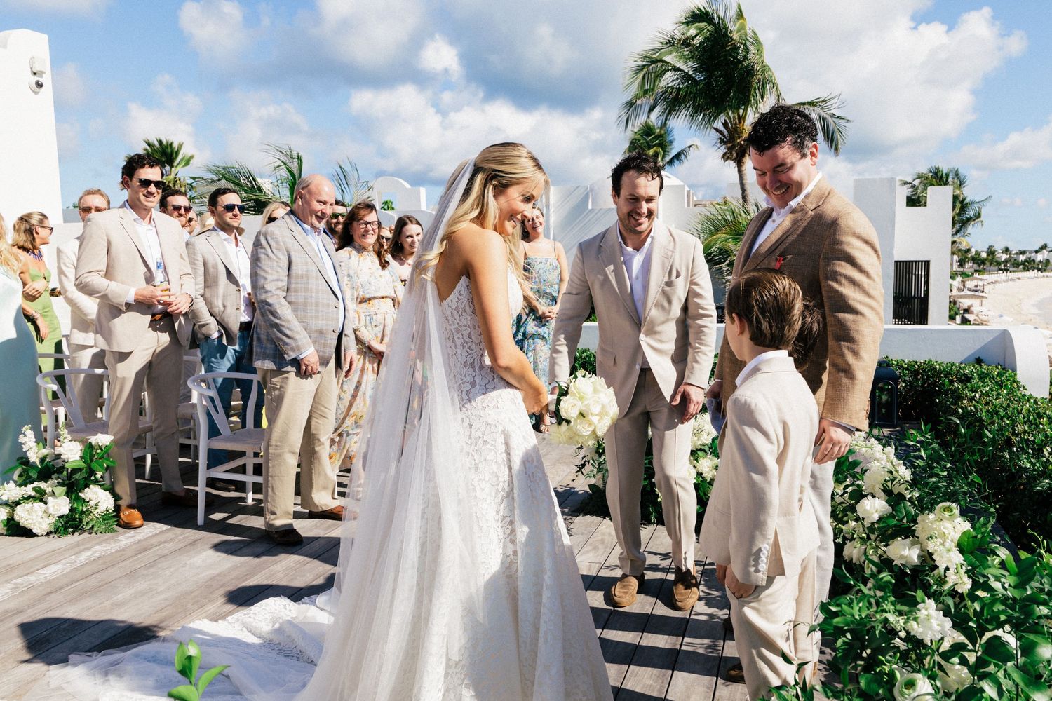 Wedding party gathered on sunny terrace with tropical landscaping and white architecture.