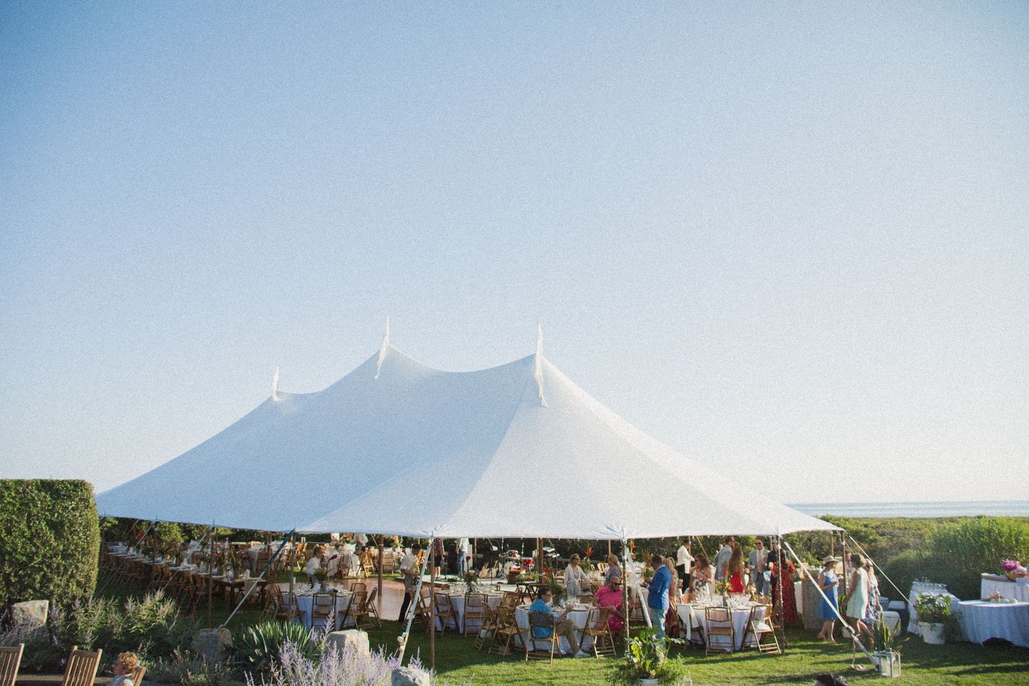 Large white wedding tent set up outdoors with guests gathering underneath.