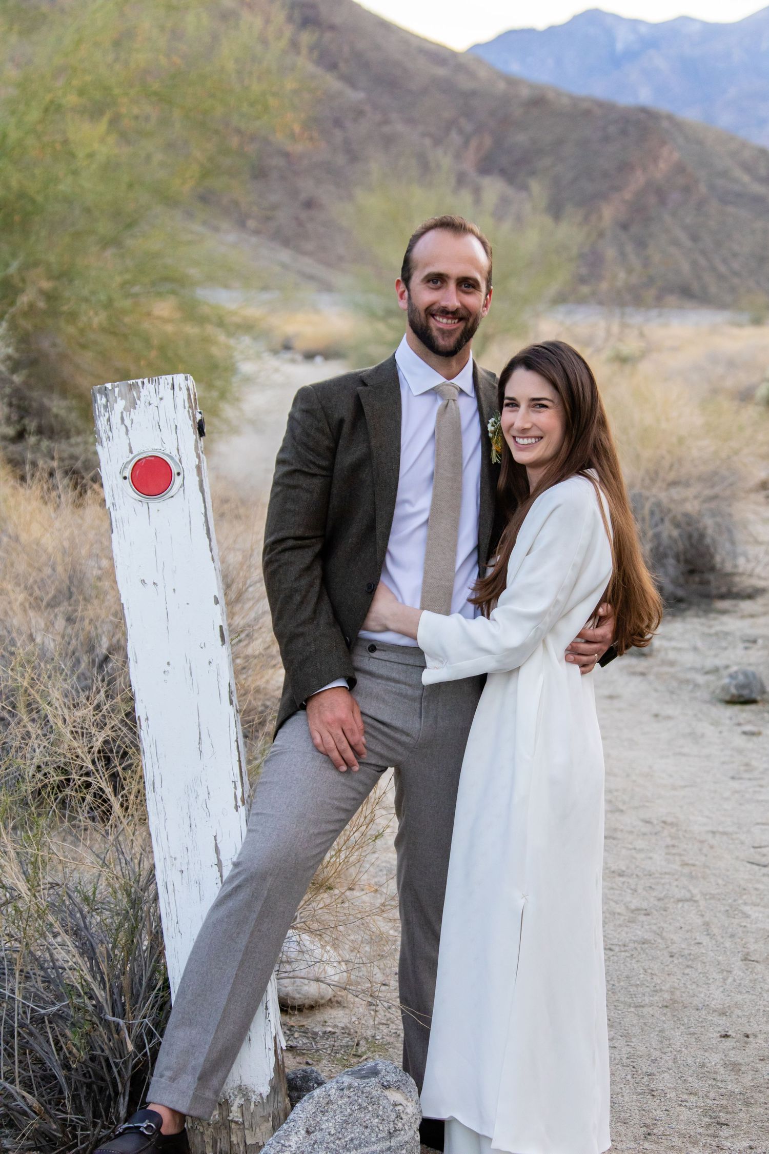 Wedding couple poses against a rustic white door in a desert setting with mountains.