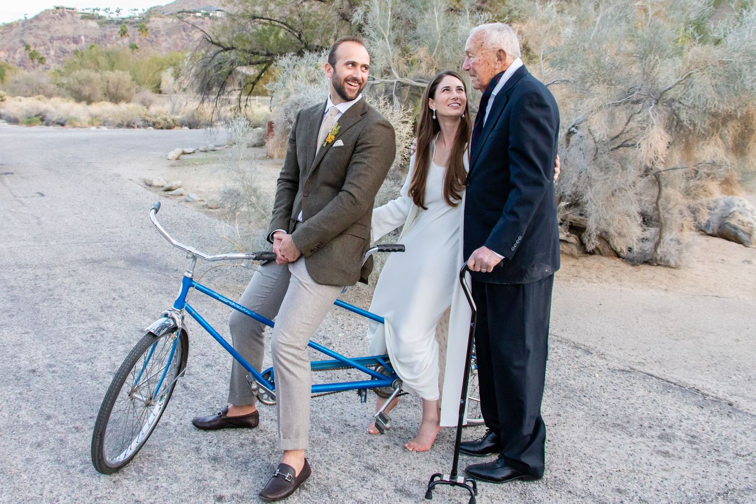 People gather around a blue bicycle in a desert setting with mountains and cacti.