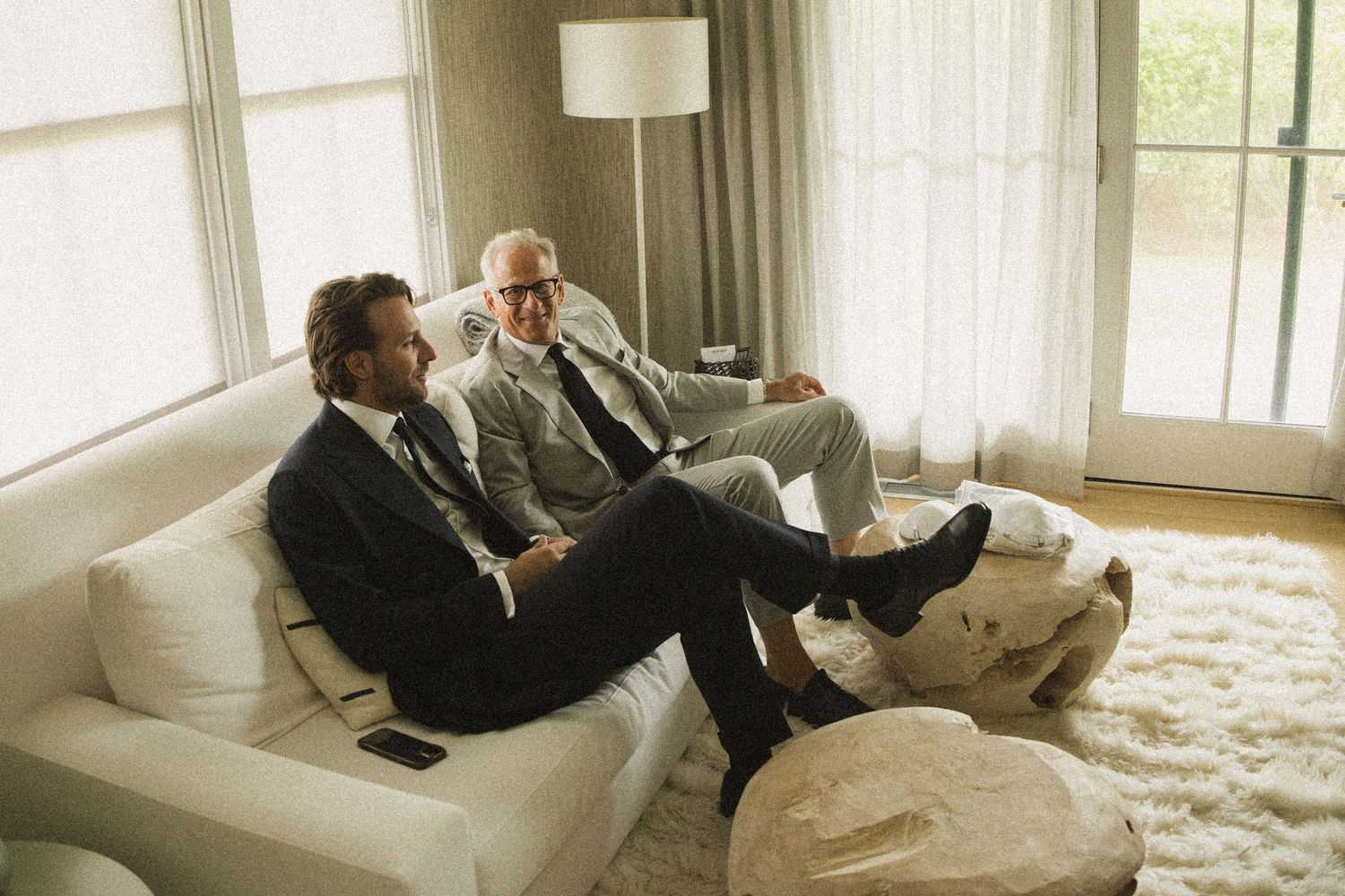 Two well-dressed men in suits relaxing on a white sofa in a bright, modern living room.