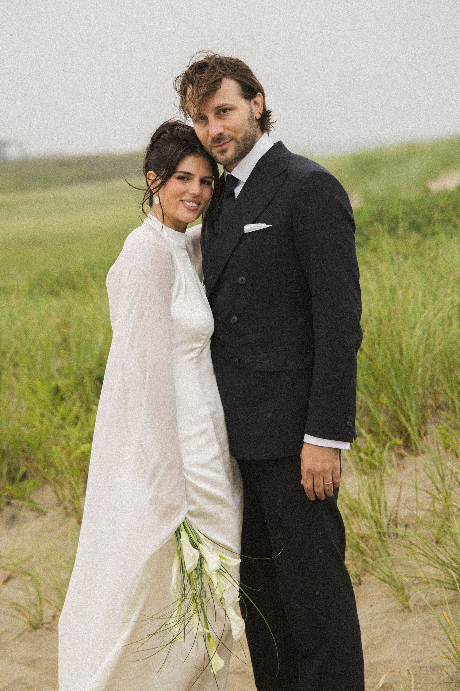Bride in white gown and groom in dark suit pose together on sandy beach with green grass backdrop.
