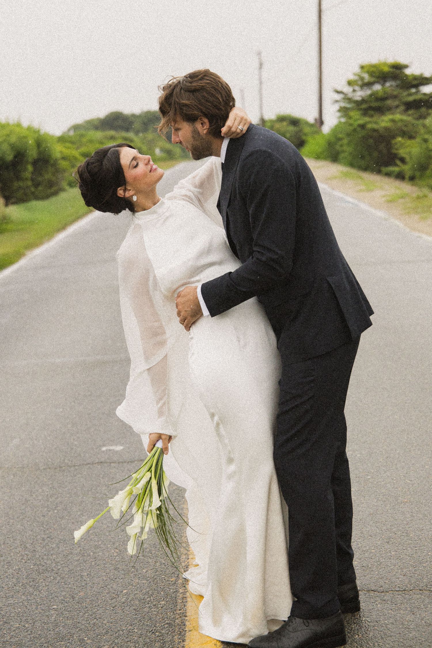 Bride in white gown holding calla lilies embraces groom in black suit on an open road surrounded by greenery.