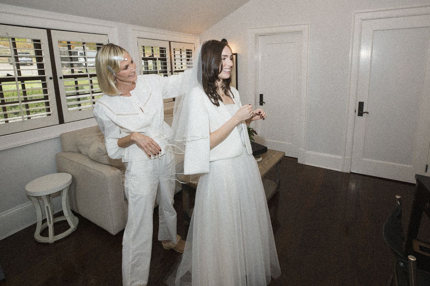 Woman in white adjusts the sleeve of a bride in a white tulle skirt and jacket inside a bright, sunlit room with a cozy armchair.