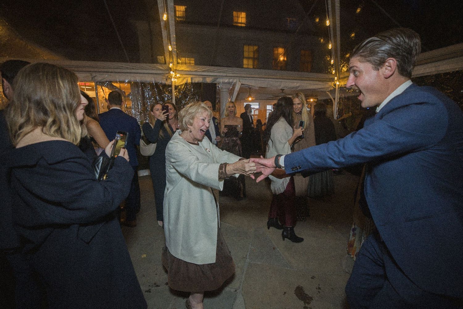 A smiling man in a navy suit dances hand in hand with an older woman in a light cardigan as guests celebrate at an evening outdoor party.