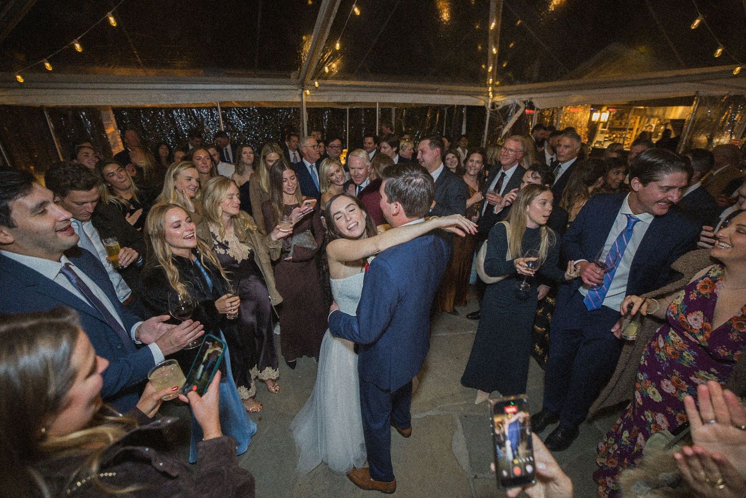 Bride and groom share a joyful dance surrounded by cheering wedding guests under a string-lit tent at an evening reception.