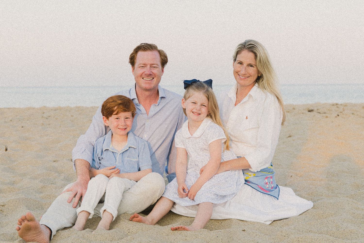 Family of four smiling together on a sandy beach at sunset, with two young children sitting on parents laps.