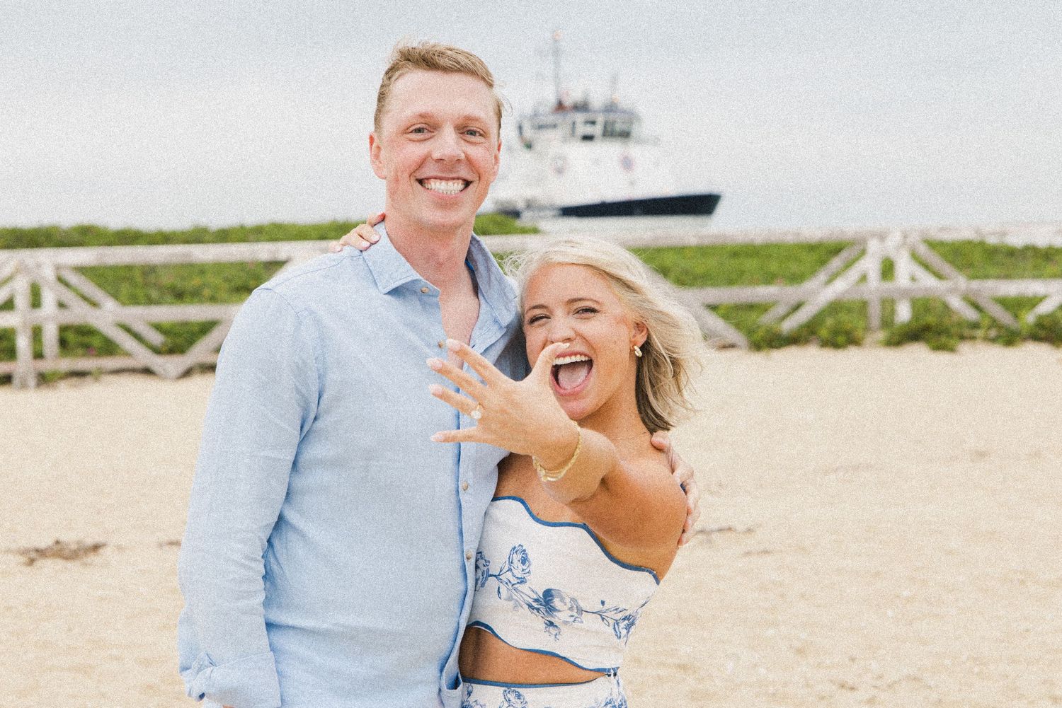 Happy couple on beach showing engagement ring, with a ship in the background on a cloudy day.