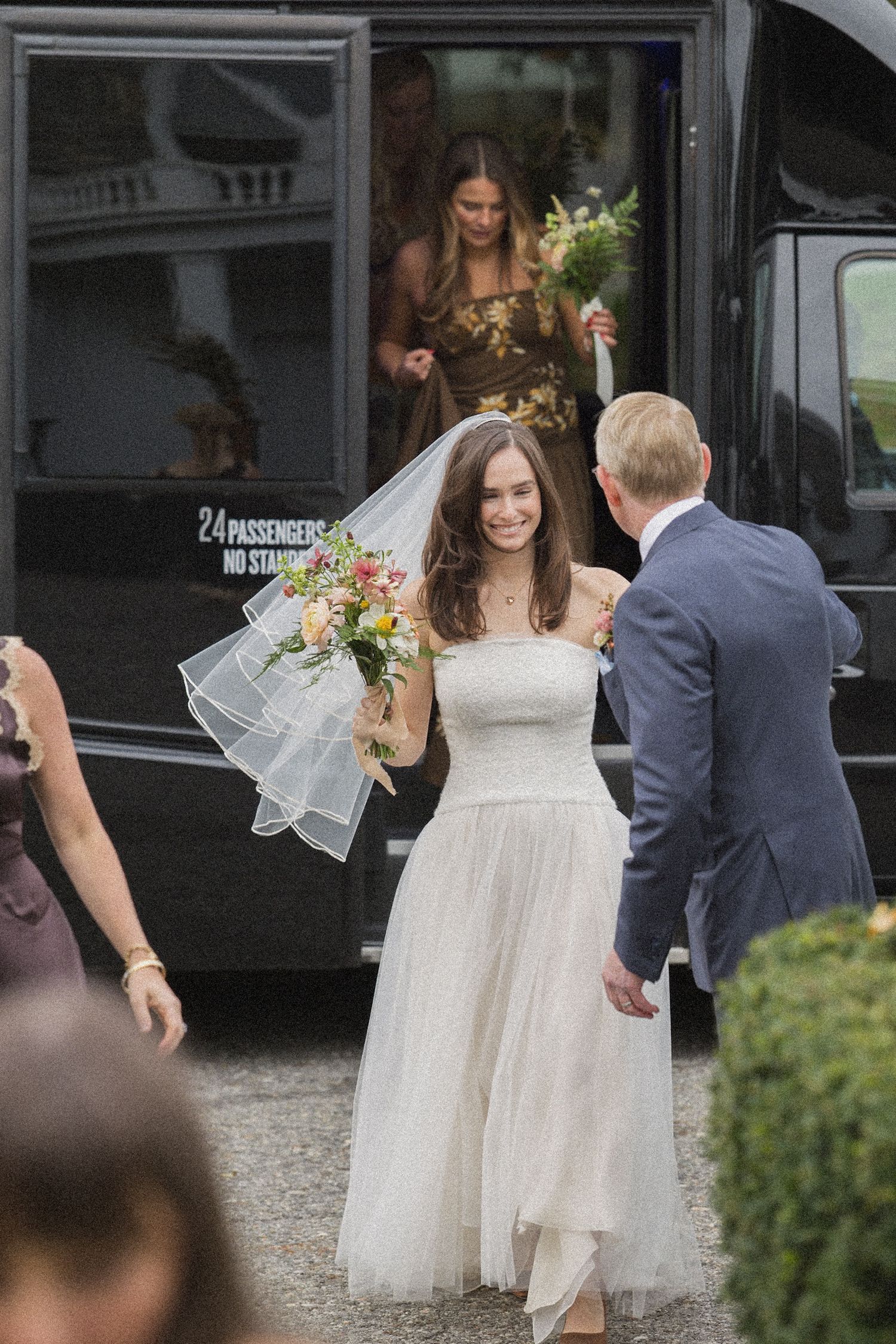 Bride in a strapless tulle gown and veil carries a colorful bouquet while stepping off a black shuttle, greeted by a man in a suit.