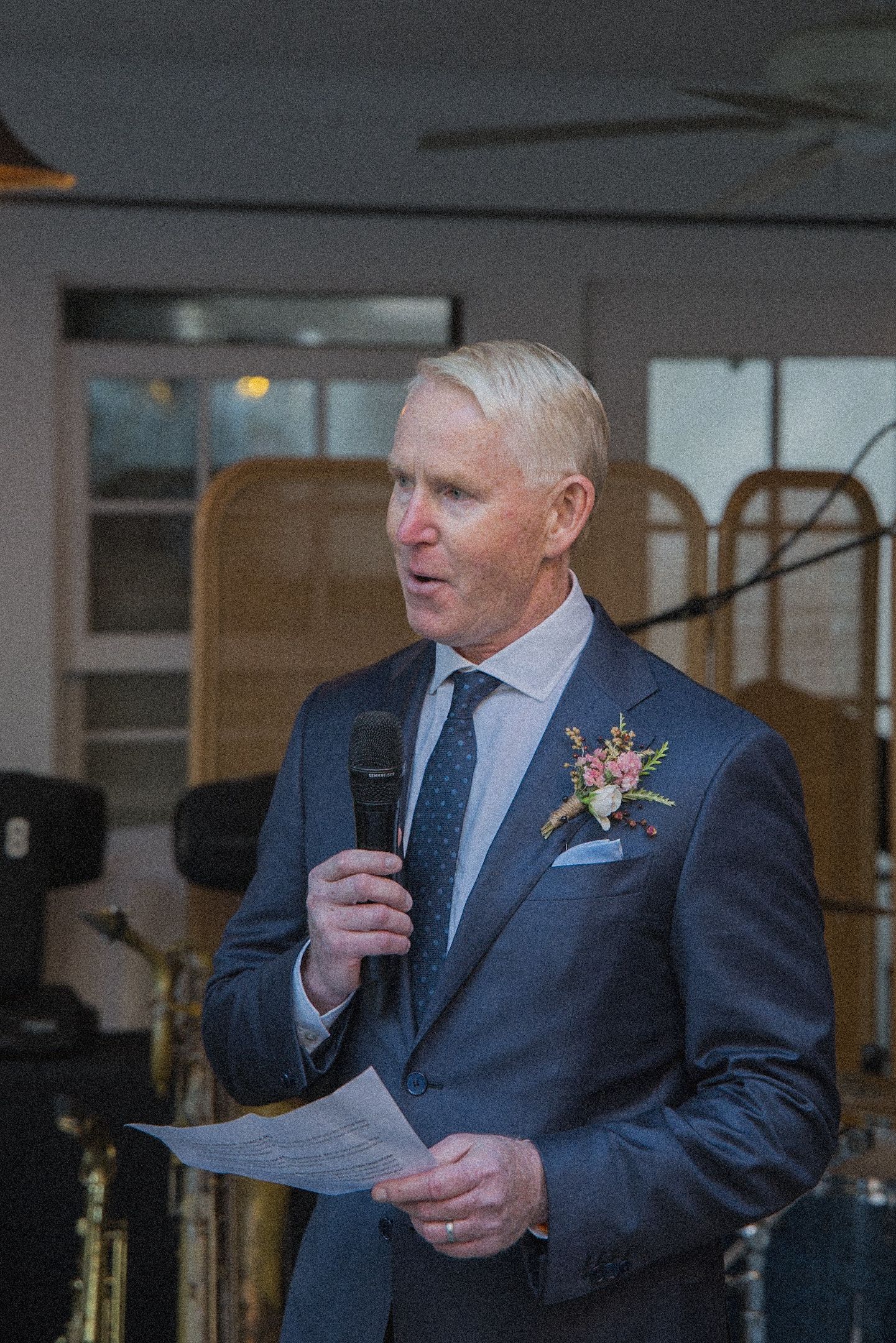 Man in a navy suit with a floral boutonniere holds a microphone and printed notes while giving a speech at an indoor event.