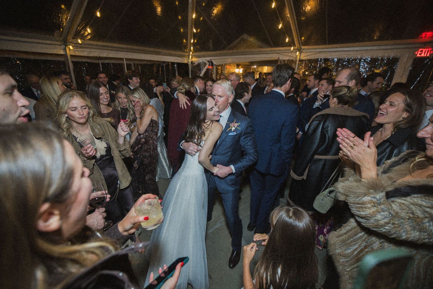 Bride in a white gown and groom in a navy suit dance closely as cheering wedding guests surround them at an evening reception under a tent.