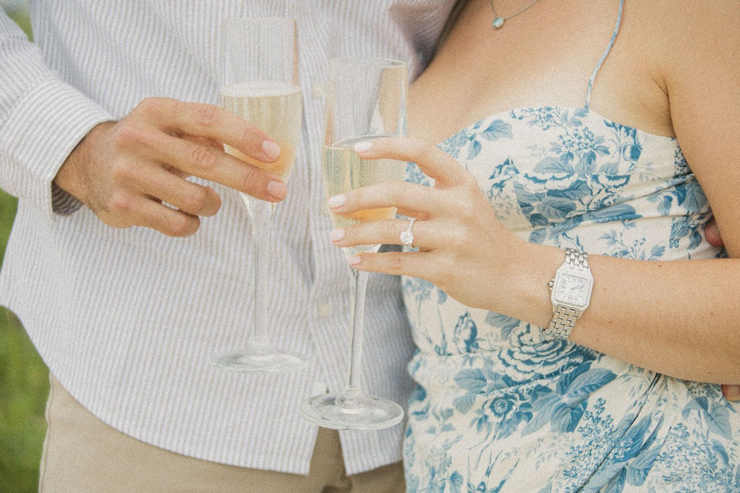 Couple toasting with champagne flutes at outdoor celebration, woman wearing blue floral dress and silver watch.