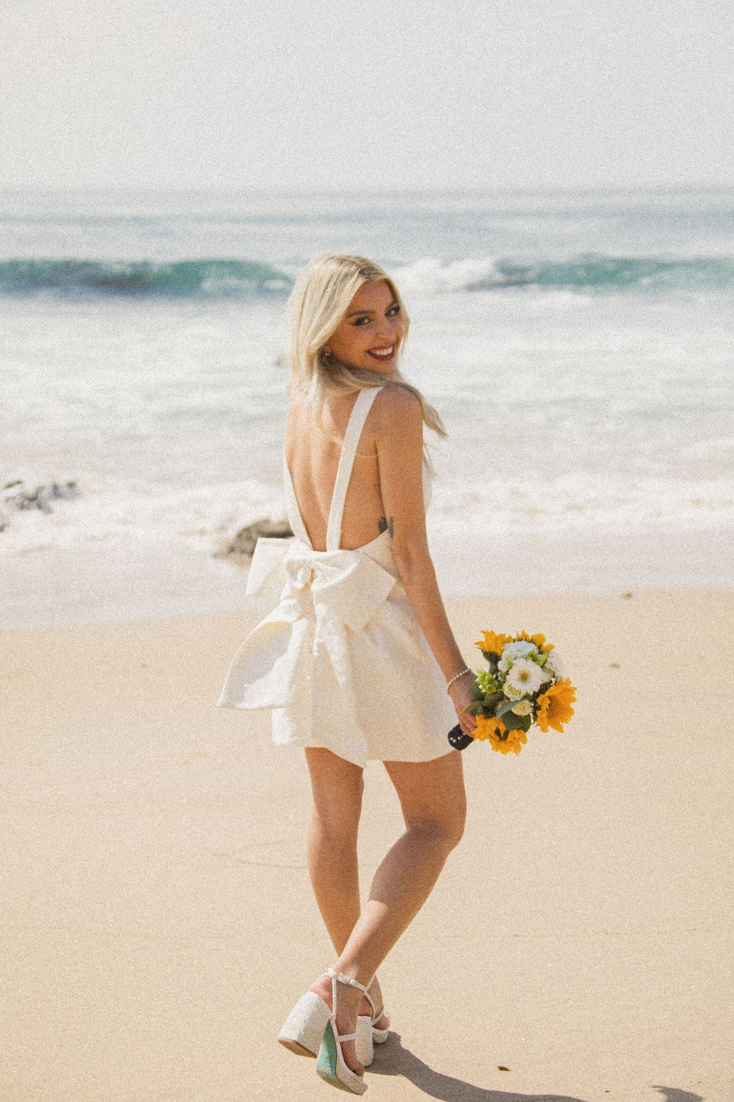 Woman in white swimsuit holding sunflowers walks along beach shoreline.