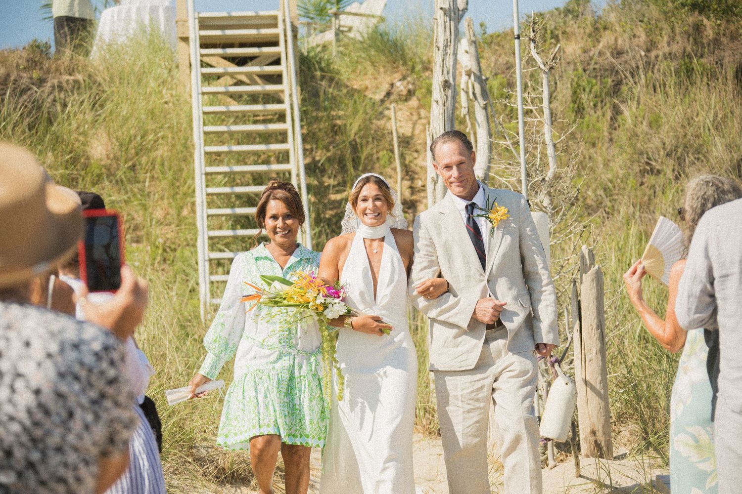 Wedding party and guests gather on sandy beach pathway during outdoor ceremony.