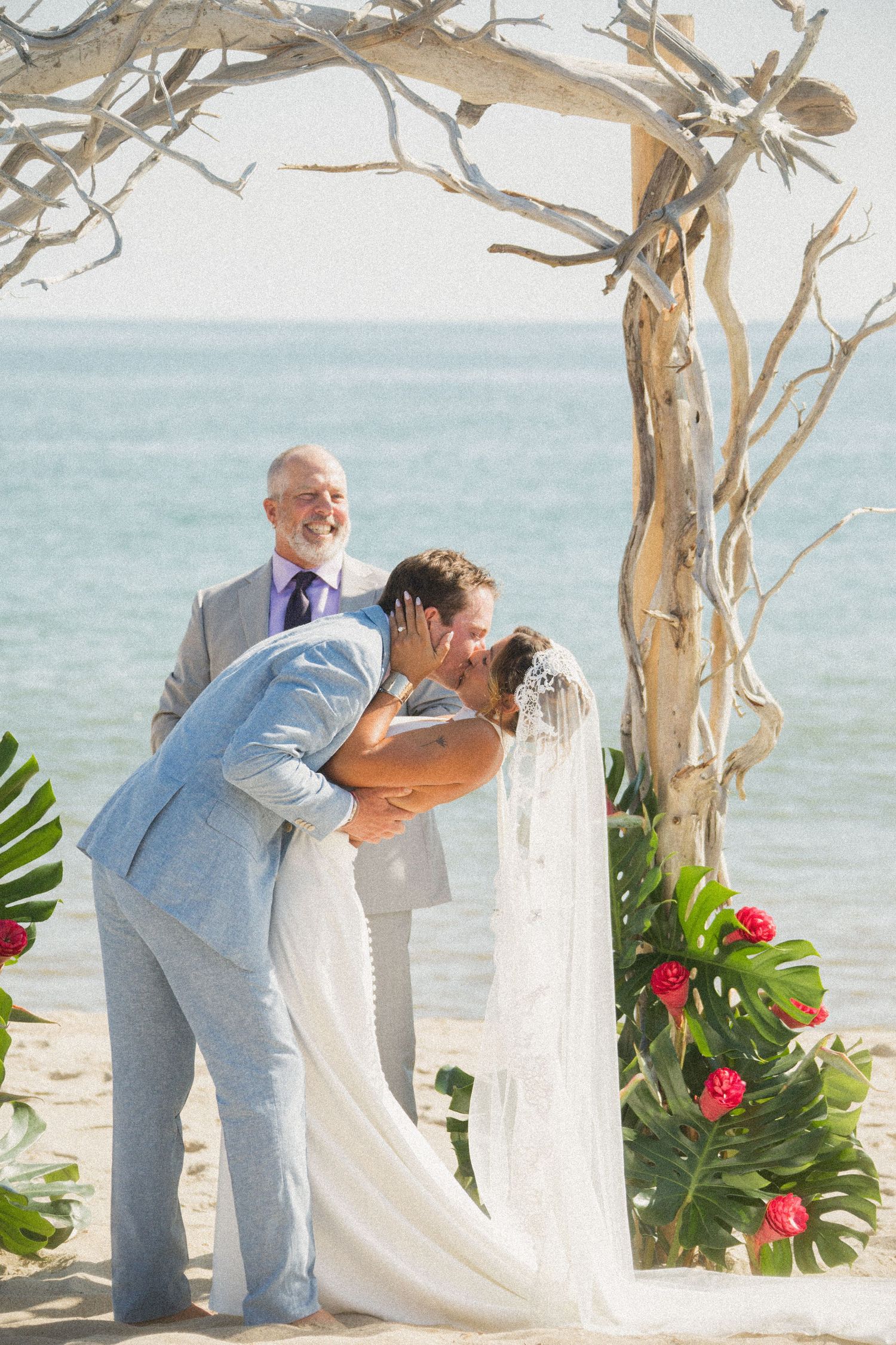 First kiss moment during beach wedding ceremony under driftwood arch.