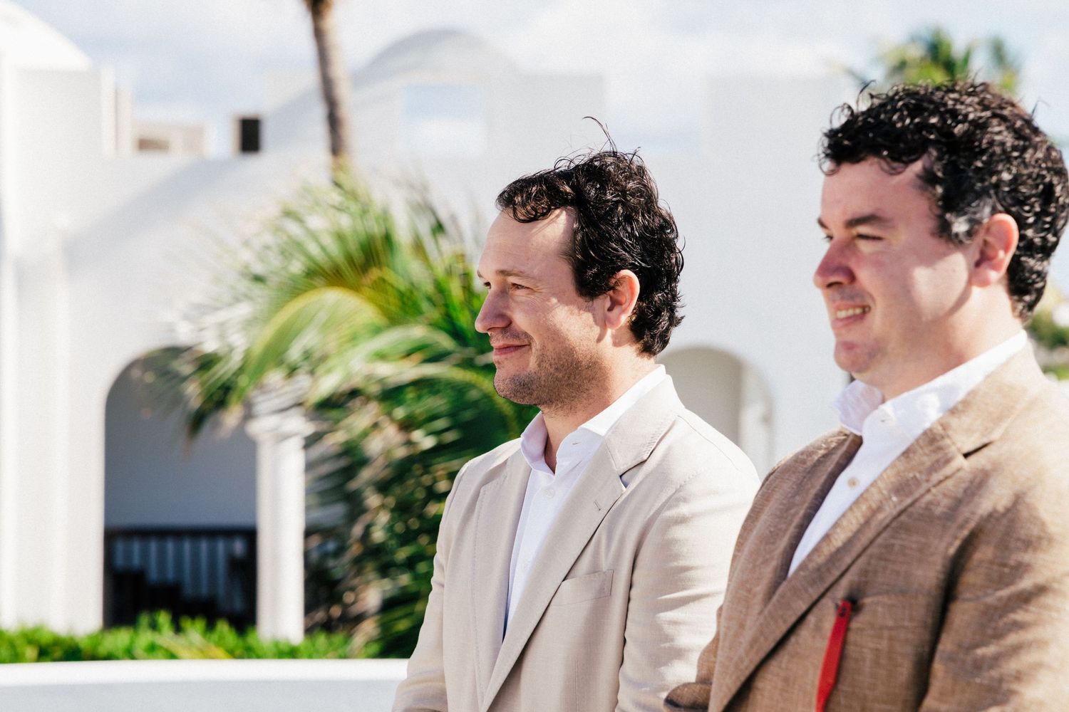 Two people in beige suits smile during outdoor ceremony with white Mediterranean architecture.