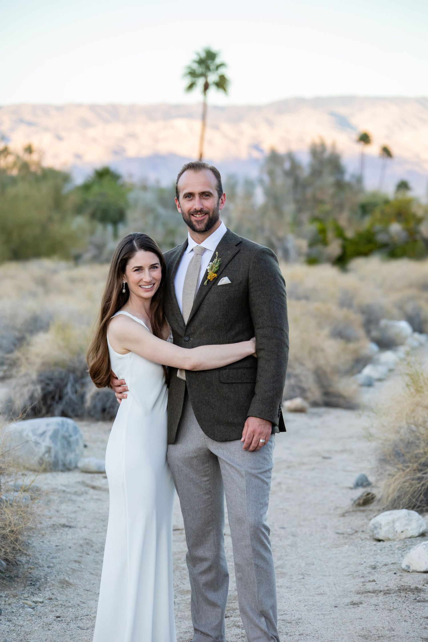 A couple poses for wedding photos in a desert landscape with mountains and palm trees in the background.
