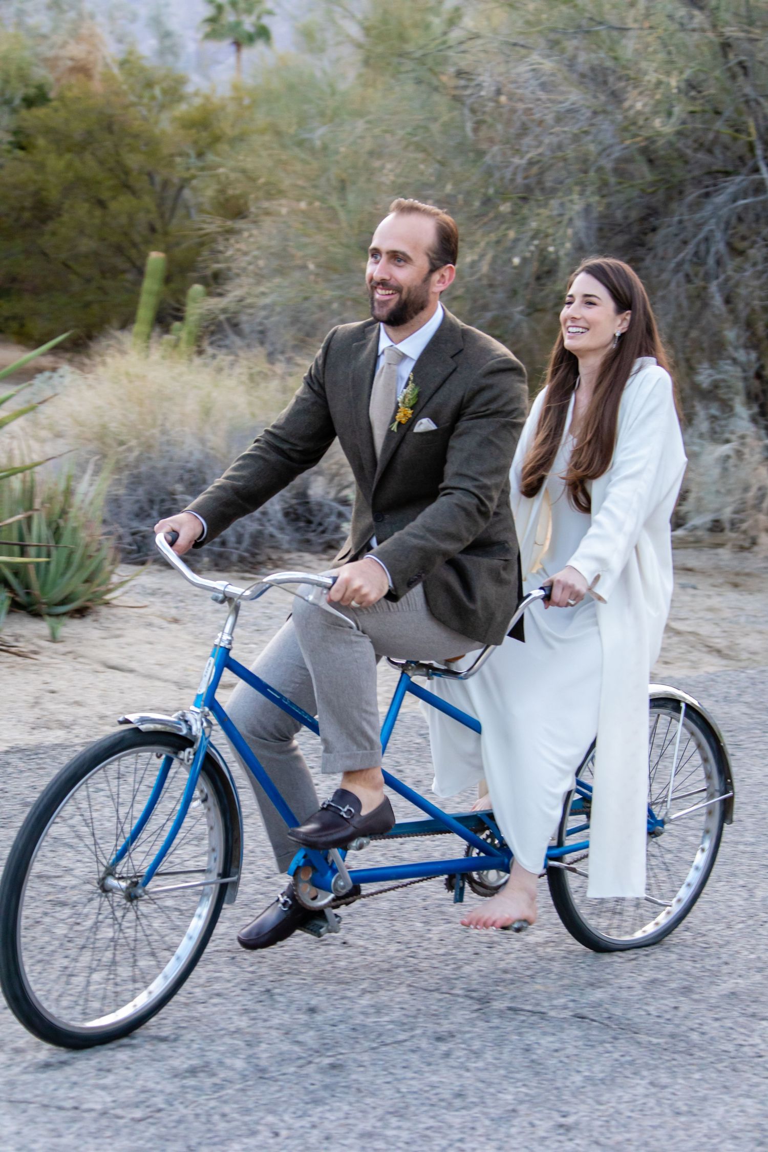 A couple rides a blue bicycle together on a desert path surrounded by desert vegetation.