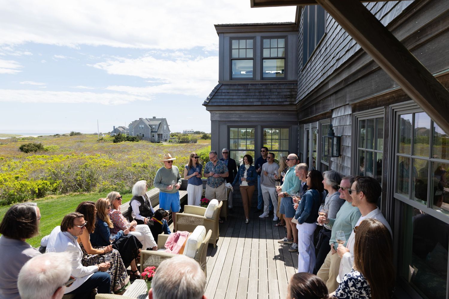Group of people gathered on a seaside deck of a modern gray shingled home on a sunny day.