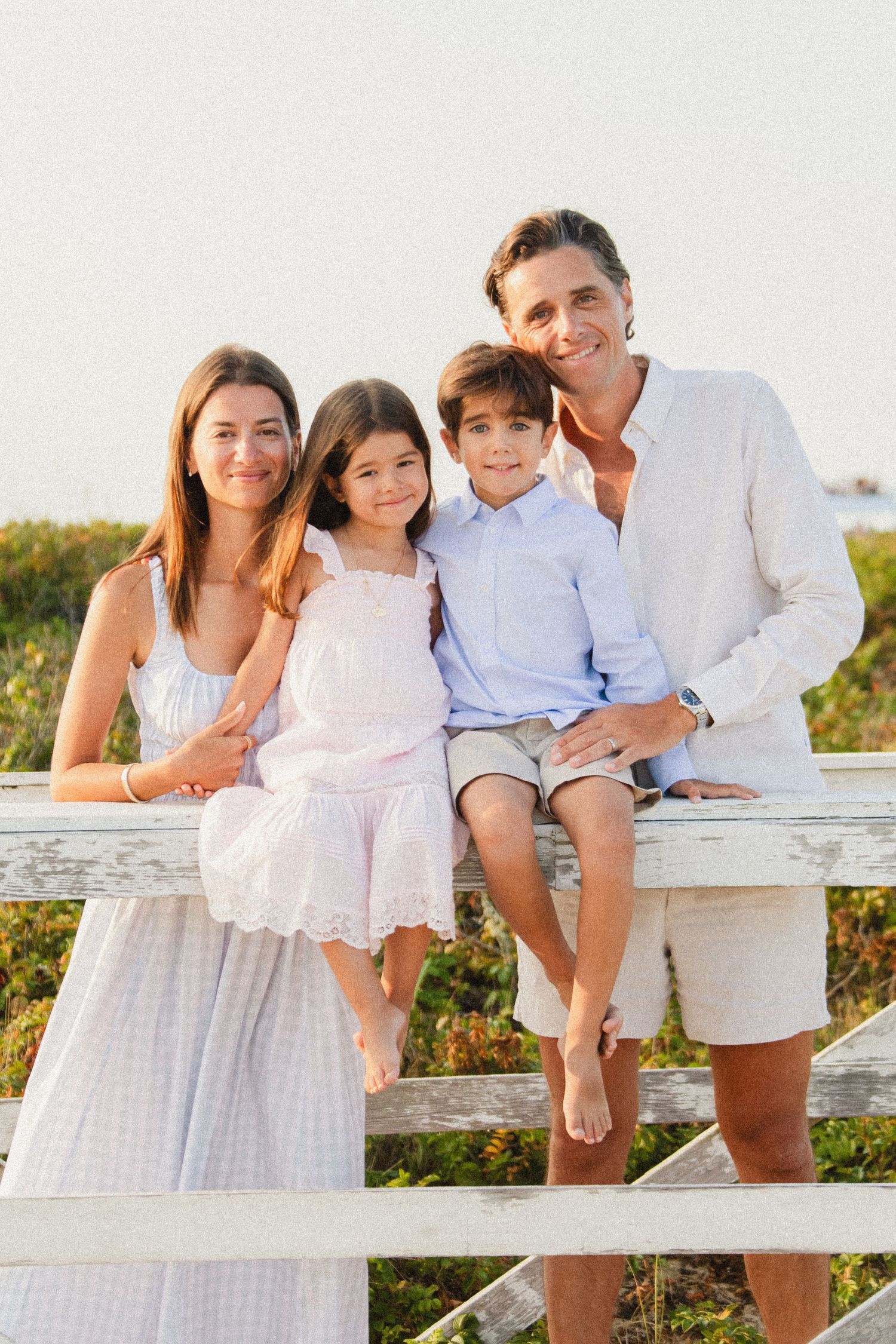 Happy family of four posing together on a white fence at the beach during golden hour.