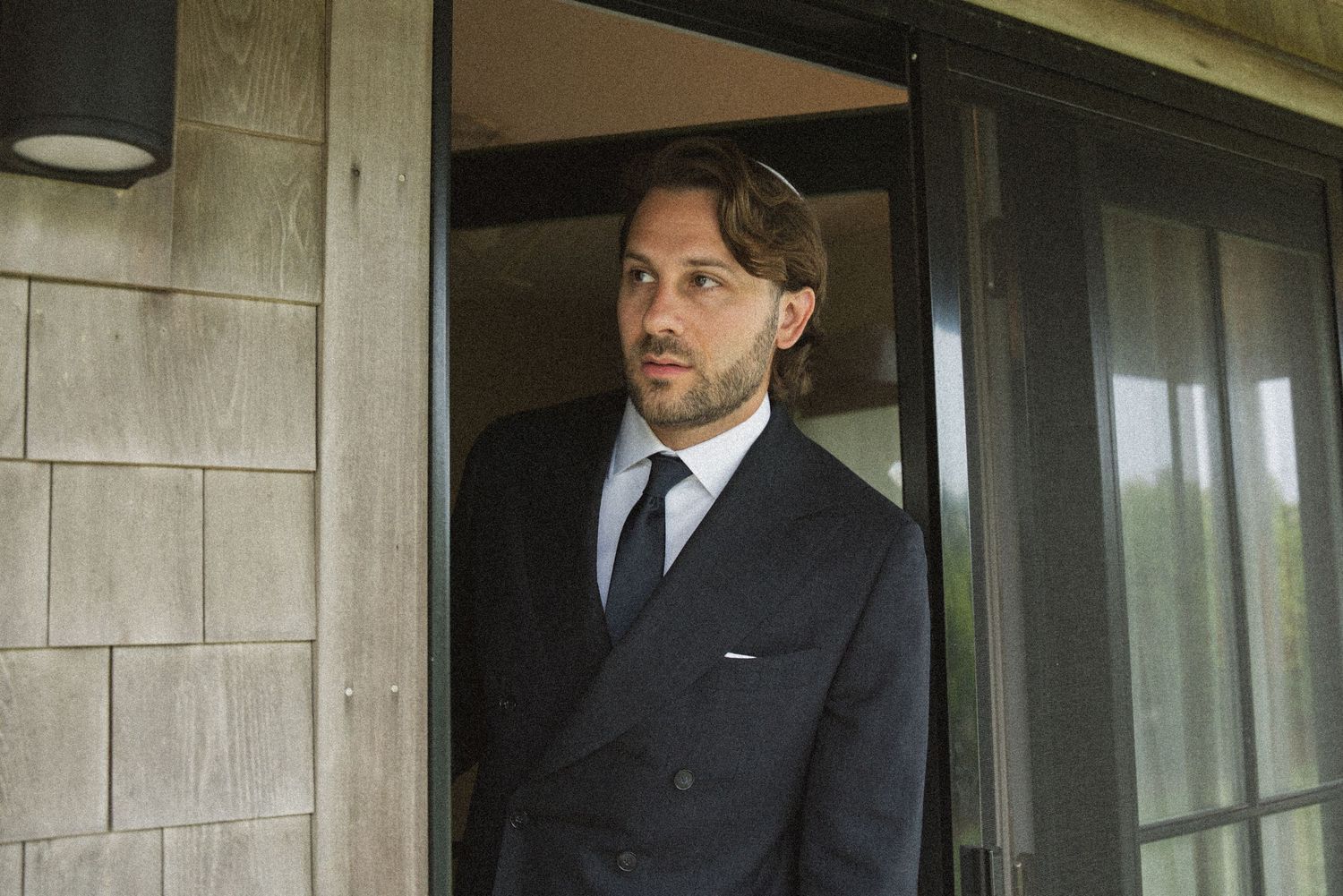 Man in dark suit and tie standing in doorway of a modern building, looking thoughtfully to the side.