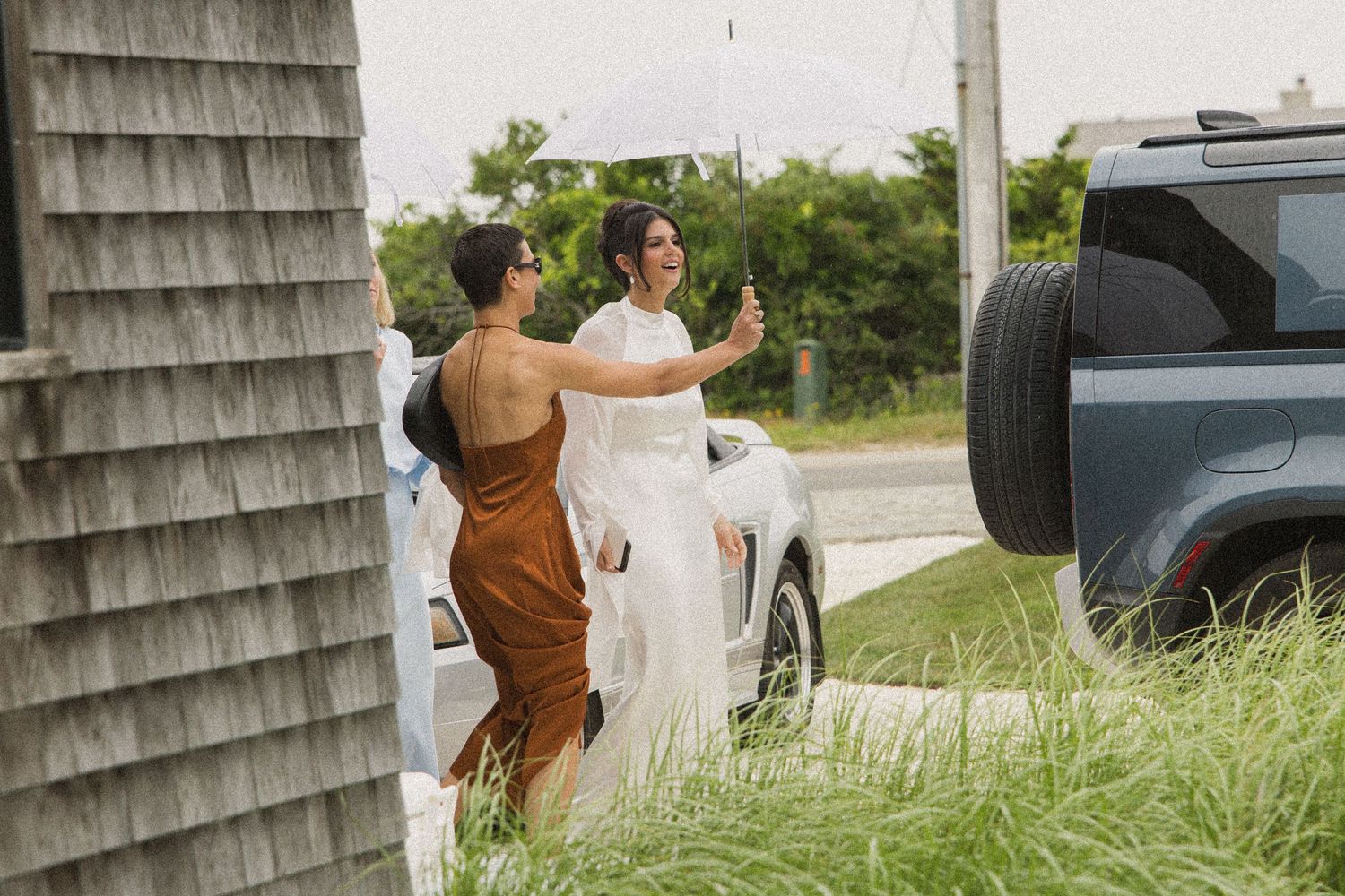 Bride in white gown greeting a guest in brown dress outdoors near a blue SUV and shingled building.
