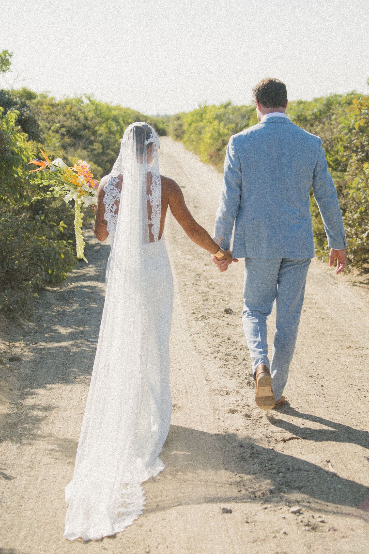 A bride in white and groom in light blue walk hand in hand down a winding coastal path.