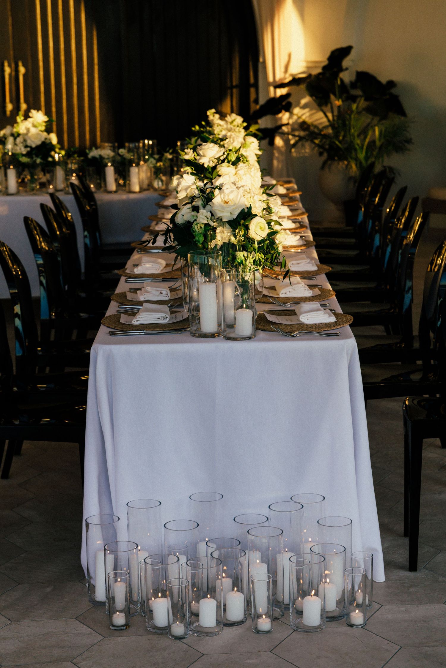 Long wedding table with white linens, candles and floral arrangements in an indoor venue.